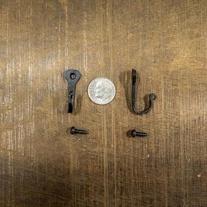 Puede incluir: Dos ganchos de metal negro con la parte superior redondeada y un solo agujero para tornillo. Los ganchos se muestran con dos tornillos negros.