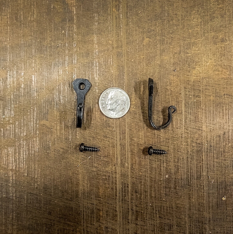 Puede incluir: Dos ganchos de metal negro con la parte superior redondeada y un solo agujero para tornillo. Los ganchos se muestran con dos tornillos negros.