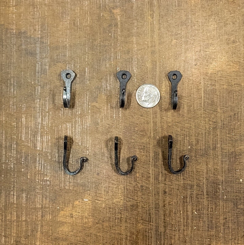 Puede incluir: Seis ganchos de metal negro montados en una superficie de madera. Los ganchos est&aacute;n en dos filas de tres. Una moneda de diez centavos de d&oacute;lar estadounidense se coloca en el centro de la imagen para la escala.