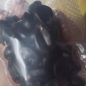 May include: A clear plastic bag filled with dark purple grapes. The grapes are tightly packed together, and the bag is slightly transparent, allowing some light to pass through. The image is taken from a close-up perspective.