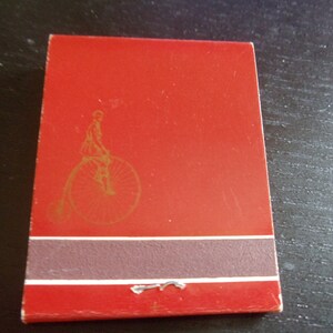 May include: Red matchbook cover with a gold illustration of a person riding a bicycle. The matchbook has a brown stripe and a white stripe.
