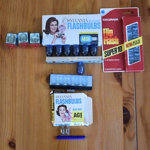 May include: A collection of vintage Sylvania flashbulbs in blue and white boxes. The boxes feature a woman holding a camera and the text "Sylvania Flashbulbs". The flashbulbs are in a variety of sizes and are labeled with the model number "M3B" and "AG1".