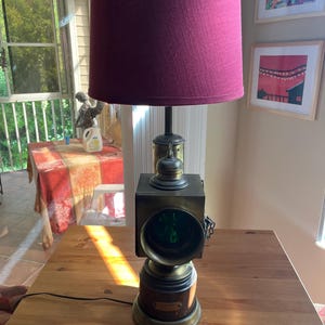 May include: An antique brass and bronze lamp with a burgundy lampshade. The lamp features a square, box-like light fixture with a green lens and a round base. A black power cord extends from the base.