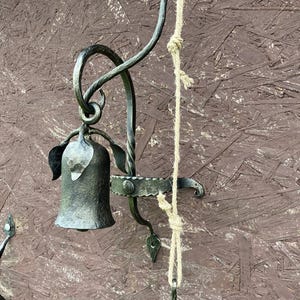 Può includere: Un'applique da parete in metallo scuro forgiata a mano, con un portacandele a forma di campana e dettagli decorativi a forma di foglia. Una corda naturale è attaccata per appenderla. L'applique ha un aspetto rustico e antico.
