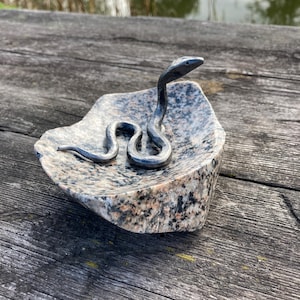 Puede incluir: Una escultura de serpiente de metal gris oscuro está fijada en el centro de un cuenco de piedra de granito pulido y moteado. La piedra presenta una mezcla de motas minerales de color tostado, negro y blanco, con una forma curva y lisa.