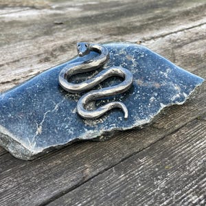 Op de afbeelding: Een handgesmeed metalen slangenbeeldje met een gehamerd, getextureerd oppervlak en een opgerold lichaam. De slang is gemaakt van donker, gepolijst staal en heeft een duidelijke kop met kleine, donkere ogen, rustend op een platte, donkere, grillige steen.