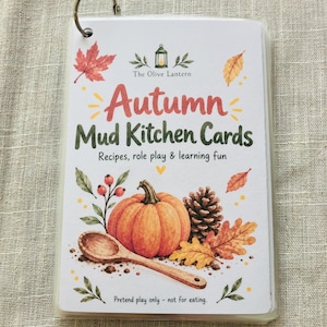 以下が含まれることがあります： 金属リングで綴じられたAutumn Mud Kitchen Cardsのセット。表紙にはカボチャ、松ぼっくり、木のスプーン、秋の葉が描かれており、The Olive Lantern, Autumn Mud Kitchen Cards, Recipes, role play & learning fun, Pretend play only - not for eating.というテキストが記載されています。
