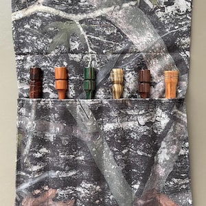 May include: A camouflage fabric case holds six wooden game calls in various shades of brown, orange, and green. The calls are cylindrical with flared ends and are arranged in a row. The case is made of a camouflage pattern fabric.