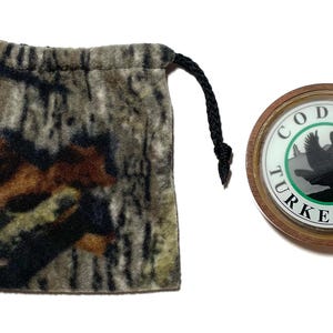 May include: A camouflage drawstring pouch and a wooden turkey call. The pouch is made of a soft, textured fabric with a brown, green, and black pattern. The turkey call has a circular design with the words "Cody Turkey Calls" and a turkey silhouette.