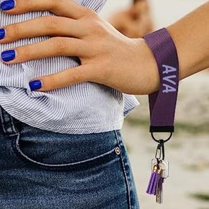May include: A purple wrist lanyard with the name "AVA" printed in white. The lanyard is attached to a keyring with keys and a purple tassel. The person is wearing a blue and white striped shirt and blue jeans.