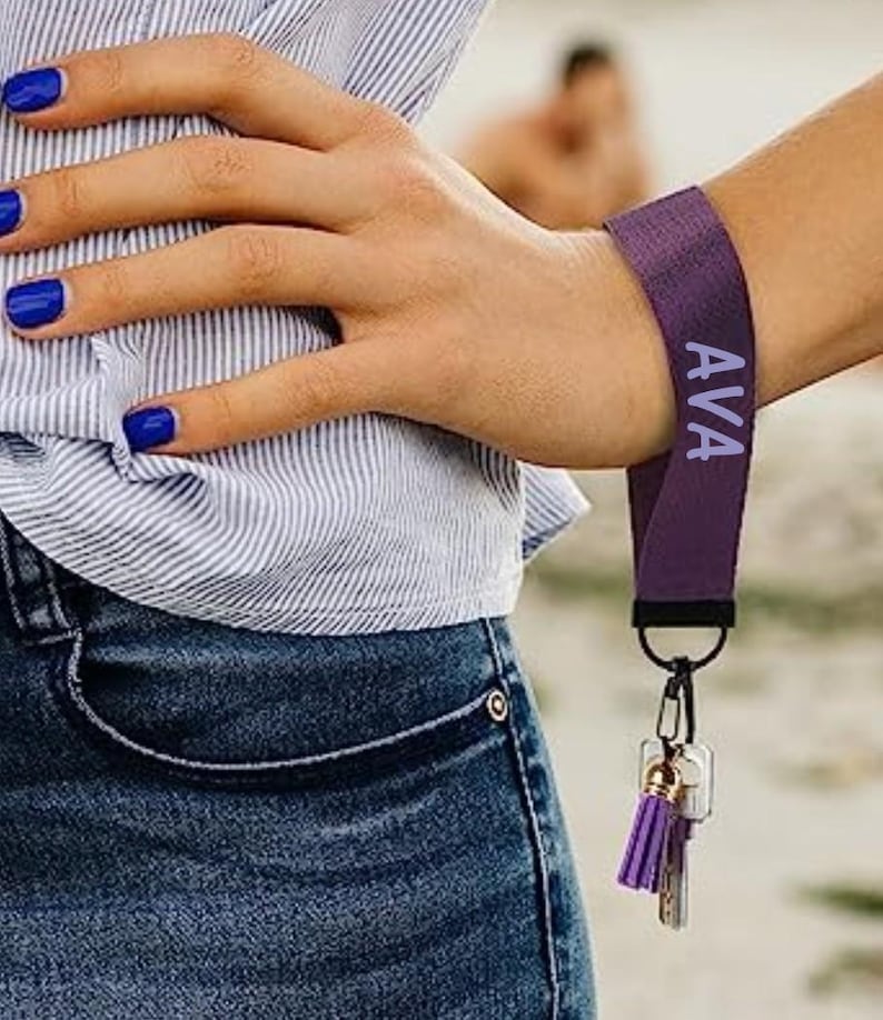 May include: A purple wrist lanyard with the name "AVA" printed in white. The lanyard is attached to a keyring with keys and a purple tassel. The person is wearing a blue and white striped shirt and blue jeans.