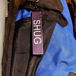 May include: A purple fabric tag with the word "SHUG" in white capital letters hangs from a black metal clip. The tag is attached to a black and blue backpack.