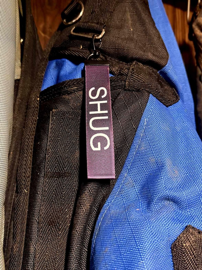 May include: A purple fabric tag with the word "SHUG" in white capital letters hangs from a black metal clip. The tag is attached to a black and blue backpack.