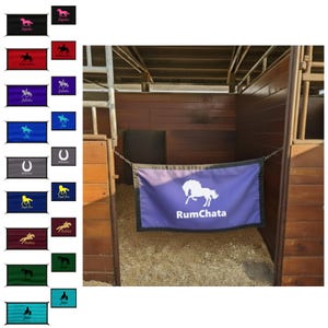 May include: A rectangular purple stall guard featuring a white graphic of a horse and the text RumChata in white lettering. The guard is bordered by a black trim and is suspended by metal chains across the opening of a wooden horse stall.
