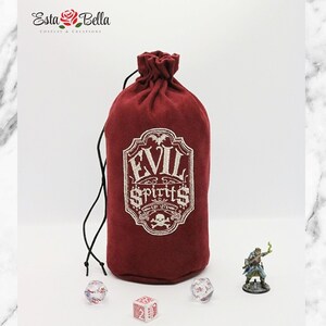 May include: A red velvet dice bag with a white embroidered design that says "Evil Spirits". The bag is tied with a black drawstring and is sitting on a white marble surface. There are three dice and a miniature figurine on the surface.