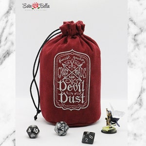May include: Red velvet dice bag with white embroidery that reads "Crossroads Devil Dust" and a white drawstring. The bag is sitting on a white marble surface with four black dice and a miniature figurine.