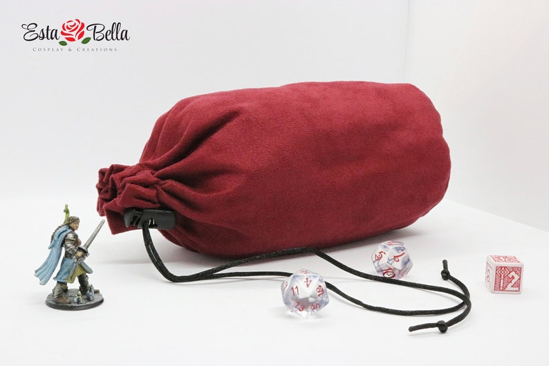 May include: A red velvet dice bag with a black drawstring and a plastic clasp. The bag is lying on a white surface with a miniature figure and dice nearby.