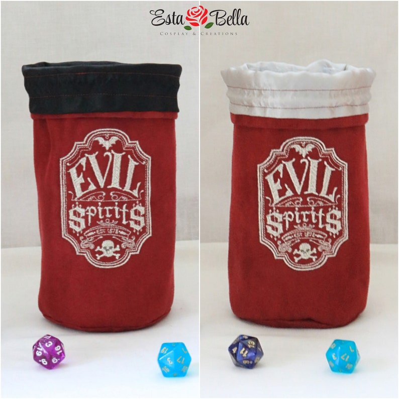 May include: Two red fabric dice bags with black and white trim. The bags have the words "EVIL SPIRITS" embroidered on them. The bags are sitting on a white surface with dice in front of them.