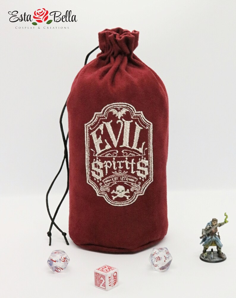 May include: A red velvet dice bag with a white embroidered design that says "Evil Spirits" with a skull and crossbones. The bag is tied with a black drawstring and is sitting on a white surface with dice and a miniature figure.