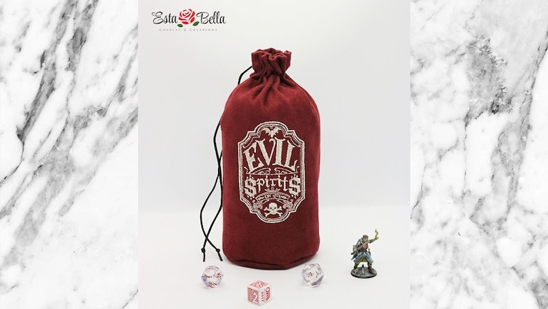 May include: A red velvet dice bag with a white embroidered design that says "Evil Spirits". The bag is tied with a black drawstring and is sitting on a white marble surface. There are three dice and a miniature figurine on the surface.