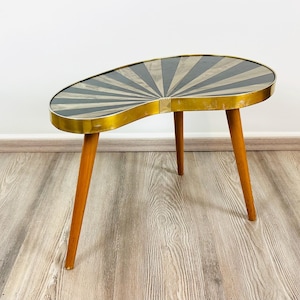 Puede incluir: Mesa auxiliar vintage en forma de riñón. La superficie presenta un diseño de rayos de sol en negro y gris, enmarcado por un borde metálico dorado. Se apoya en tres patas de madera cónicas, combinando diseño retro y funcionalidad. Mide unos 76 cm de largo.