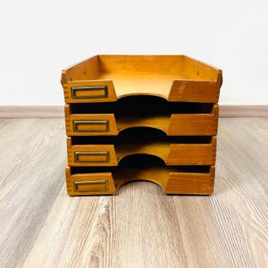 Może przedstawiać: Czteropoziomowy drewniany organizer na dokumenty w jasnobrązowym kolorze. Każda z czterech ułożonych w stos tacek posiada prostokątny uchwyt na etykiety w kolorze mosiądzu na przedniej krawędzi oraz wycięcie ułatwiające dostęp do papierów.