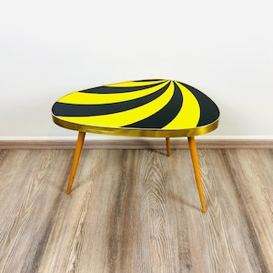 Peut inclure: Une table d'appoint de style rétro avec un design saisissant. Le plateau présente un motif en spirale noir et jaune, bordé d'un liseré doré. Elle repose sur trois pieds en bois. Dimensions: environ 70 cm de long.