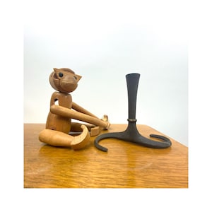 Könnte beinhalten: Ein hölzernes Affenspielzeug mit beweglichen Gliedmaßen sitzt neben einem schwarzen Kerzenhalter aus Metall mit einem dreibeinigen Fuß.