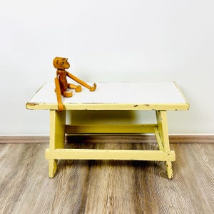 Könnte beinhalten: Kleiner, rechteckiger Tisch aus gelb und weiß lackiertem Holz mit einem Used-Look. Auf dem Tisch sitzt eine Holzfigur eines Affen. Der Tisch ist ca. 61 cm lang, 30 cm breit und 25 cm hoch.