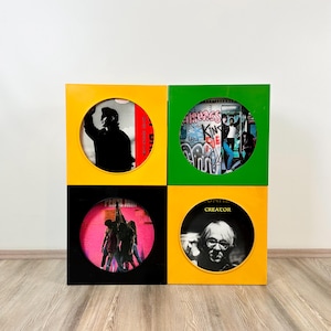Op de afbeelding: Een vinylplatenopslag met vier compartimenten. Elk compartiment heeft een andere kleur: geel, groen, zwart en geel. Elk compartiment heeft een cirkelvormige opening met een albumhoes. De unit is van hout en staat op een houten vloer.