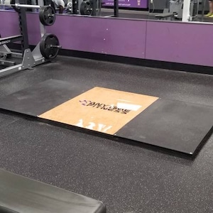 May include: A black and brown weightlifting platform with the text "Anytime Fitness" printed on it. The platform is in a gym with a purple wall and a mirror reflecting other gym equipment.