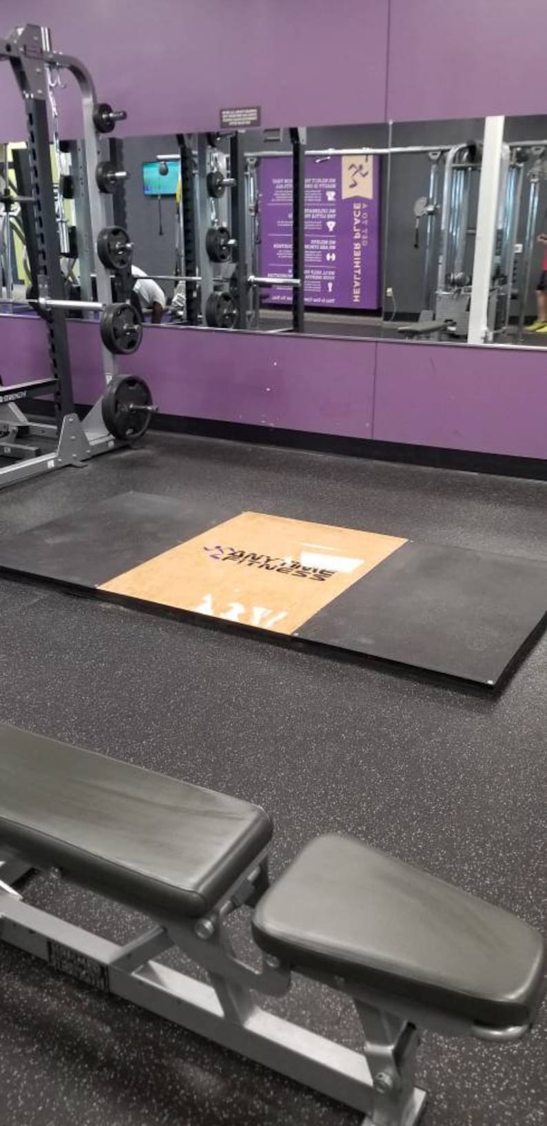 May include: A black and brown weightlifting platform with the text "Anytime Fitness" printed on it. The platform is in a gym with a purple wall and a mirror reflecting other gym equipment.
