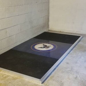 May include: A black and blue floor mat with a silver border and a navy blue seal with a white eagle and anchor in the center. The seal is surrounded by a white rope design.