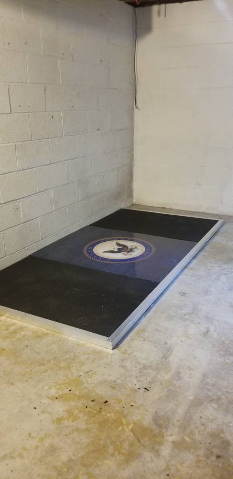 May include: A black and blue floor mat with a silver border and a navy blue seal with a white eagle and anchor in the center. The seal is surrounded by a white rope design.
