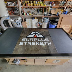 May include: A black and gray weightlifting platform with a wooden surface. The platform has a logo that says "Surplus Strength &" with a red and white triangle above the text.