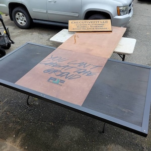May include: A black and brown platform with the text "You can't cheat the grand" written on it. The platform is sitting on two black metal folding chairs. A wooden sign with the text "ExecutiveFit LLC" is on the platform. The sign lists services offered by the company, including custom platforms, barbell storage, and epoxy inlays and signage.