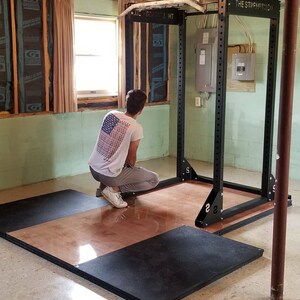 May include: A black weightlifting rack with a pull-up bar, labeled "THE STRENGTHLOD", is set up in a basement. A person is assembling the platform for the rack, which is made of black and brown rubber.