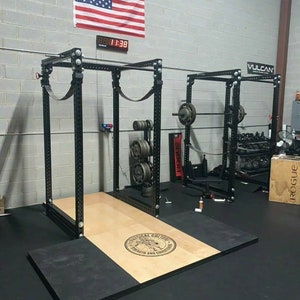 May include: Two black weightlifting racks with a wooden platform and black rubber mats. The racks have a pull-up bar, safety bars, and weight plates. The platform has a logo that says "Tactical College Strength and Conditioning".