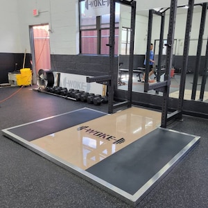 May include: A black weightlifting platform with a beige center section. The platform has a silver metal border and the words "WOKE" in black lettering are printed on the beige section.