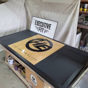 May include: A black and wood framed sign with the text "The Human Elite" and a black circle with the letters "E.F." inside. The sign is on a wooden table with a white sign that says "Executive Fit" in the background.