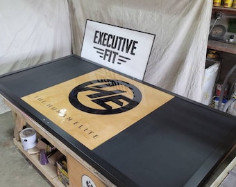 May include: A black and wood framed sign with the text "The Human Elite" and a black circle with the letters "E.F." inside. The sign is on a wooden table with a white sign that says "Executive Fit" in the background.