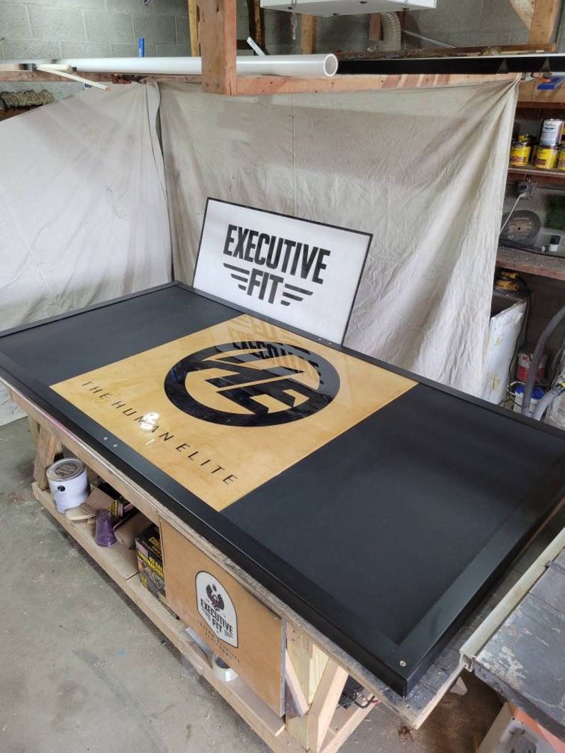 May include: A black and wood framed sign with the text "The Human Elite" and a black circle with the letters "E.F." inside. The sign is on a wooden table with a white sign that says "Executive Fit" in the background.