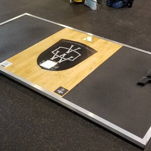 May include: A black and silver weightlifting platform with a wooden surface. The platform has a logo with the letters "WA" in a shield shape.
