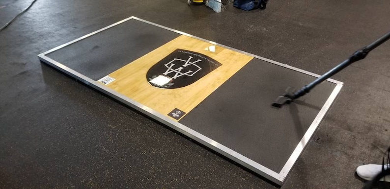 May include: A black and silver weightlifting platform with a wooden surface. The platform has a logo with the letters "WA" in a shield shape.
