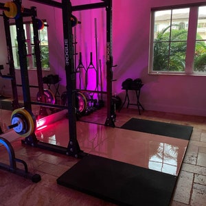 May include: A black weightlifting rack with the word "ROGUE" printed on the side. The rack is set up in a home gym with pink lighting and a black weightlifting mat on the floor.