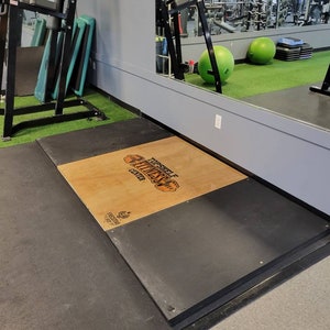 May include: A black and gray platform with a wooden top. The wooden top has the text "Top Shelf Fitness Center" printed on it. The platform is in a gym setting.