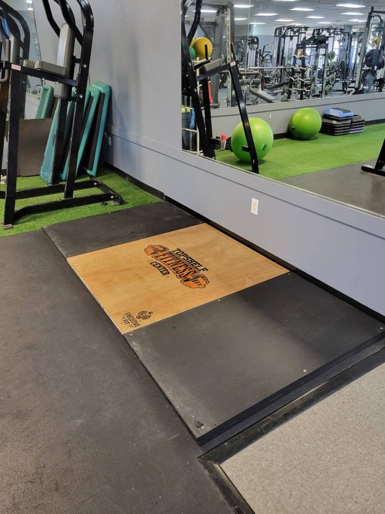 May include: A black and gray platform with a wooden top. The wooden top has the text "Top Shelf Fitness Center" printed on it. The platform is in a gym setting.