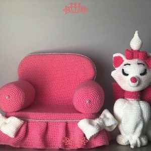 May include: A pink crocheted armchair with white trim and a white crocheted cat doll wearing a pink bow. The armchair has a ruffled skirt and the cat doll has a white bow around its neck.