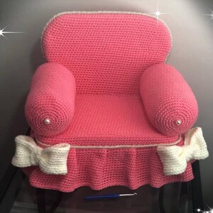 May include: A pink crocheted armchair with white bows and pearl accents. The armchair has a ruffled skirt and is made with a soft, textured yarn.