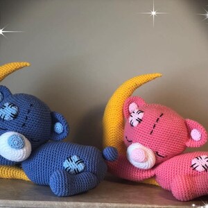 Puede incluir: Dos osos de peluche de crochet durmiendo en lunas crecientes amarillas. El oso de la izquierda es azul con la cara blanca y el oso de la derecha es rosa con la cara blanca.